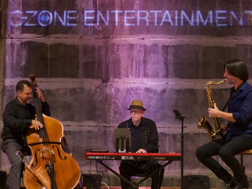 Jazz trio on upright bass, keys and sax beneath the C-Zone Entertainment sign
