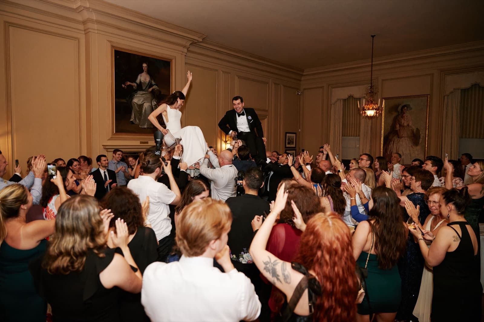 A packed dance floor in the Crane Estate ballroom, the bride lifted on a chair above the crowd