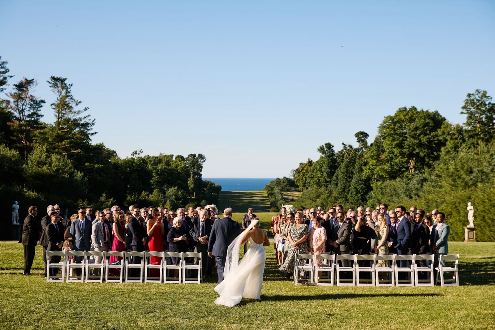A ceremony on the Grand Allée at the Crane Estate, guests standing as the bride arrives, the ocean beyond the trees