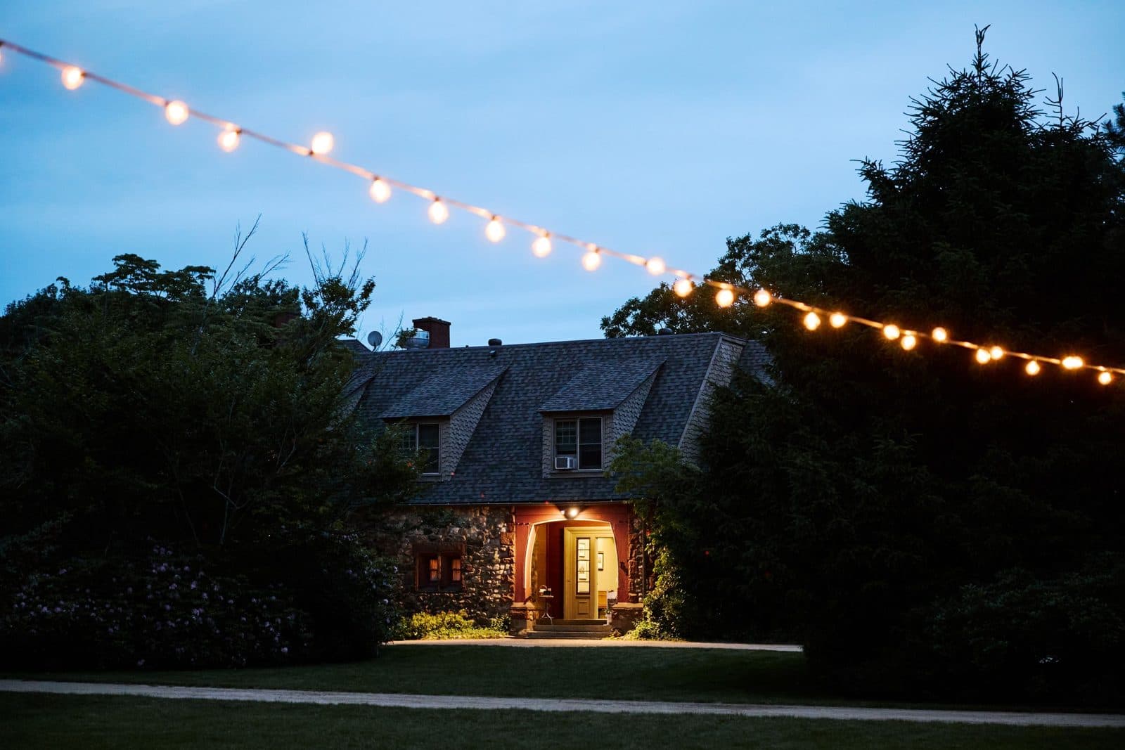 The stone house at Moraine Farm at dusk, string lights strung across the lawn