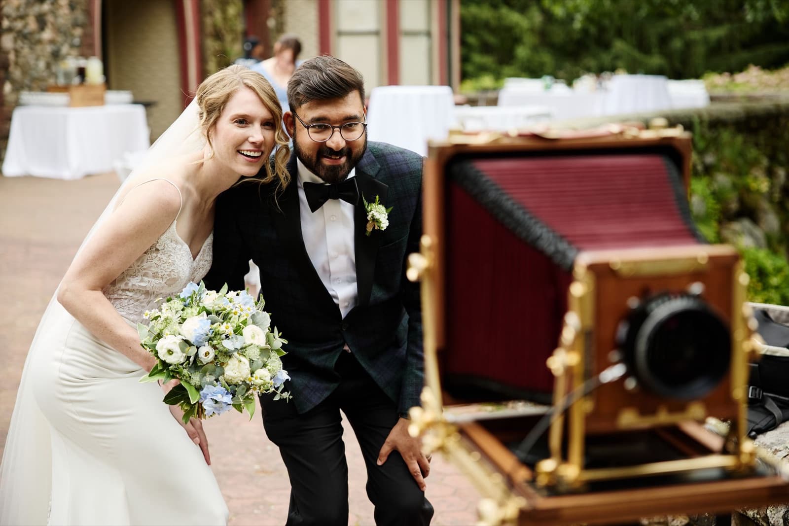 A couple posing at a vintage camera on the brick terrace at Moraine Farm, cocktail tables behind them