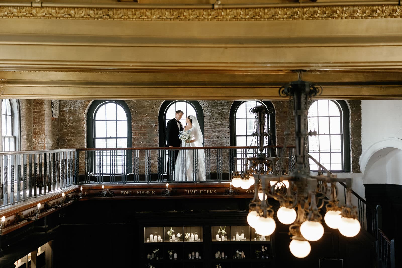 A couple on the balcony of the main hall at The Tower in Boston, under the chandeliers and the arched windows