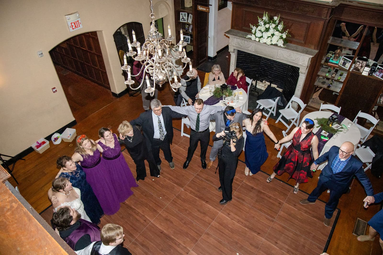 Guests dancing in a circle in the Willowdale Estate Great Room, seen from the loft, with the fireplace and chandelier