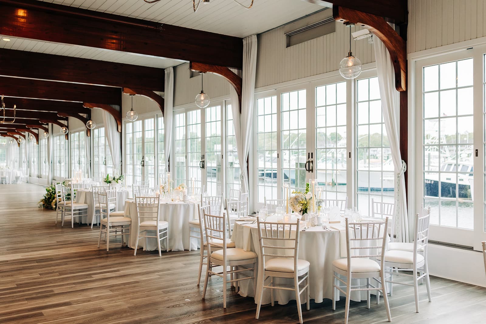 The Harbor Room at Wychmere Beach Club set for dinner, its tall windows looking out on boats in the harbor