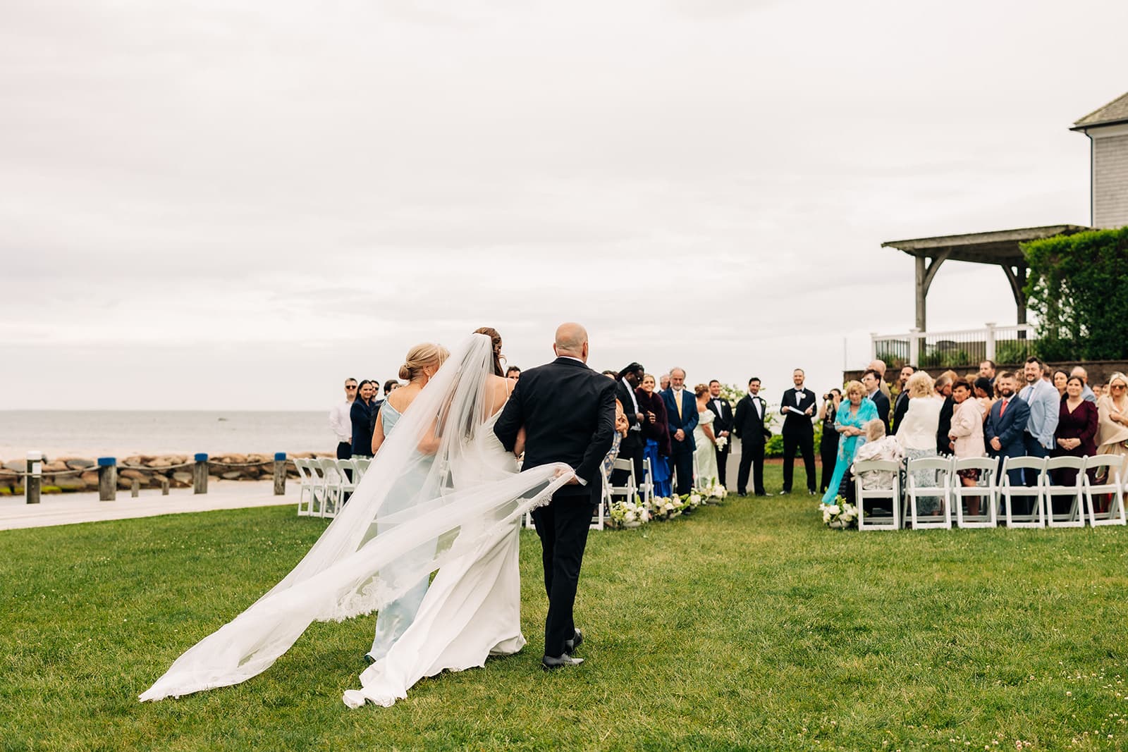 A bride walking down the aisle on the lawn at Wychmere Beach Club, guests standing and the harbor and Nantucket Sound beyond