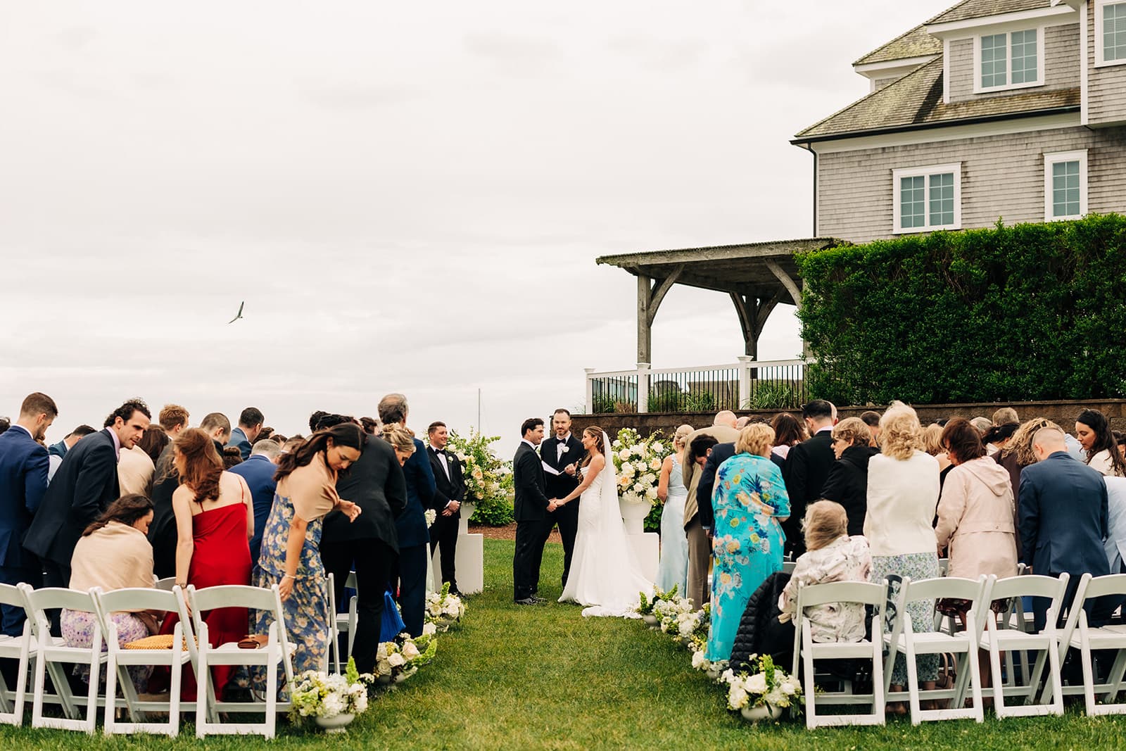 A ceremony on the lawn at Wychmere Beach Club, guests in white chairs and the pergola and shingled clubhouse behind the couple